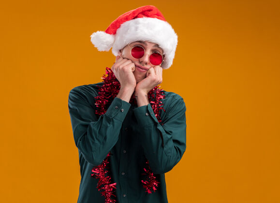 年轻无聊的金发年轻人戴着圣诞帽 戴着眼镜 脖子上戴着金箔花环 看着相机 手放在下巴上 在橙色背景上 留着复印空间眼镜金发保持