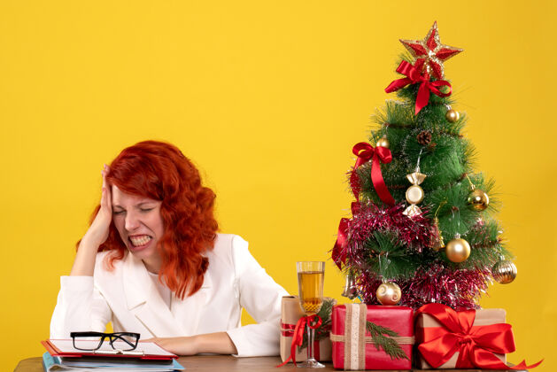 办公室女工人坐在桌子后面 拿着圣诞礼物 树在黄色上生气女性礼物桌子