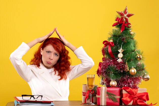 圣诞女工人坐在桌子后面 手里拿着圣诞礼物 树在黄色的树上感到悲伤桌子黄色礼物