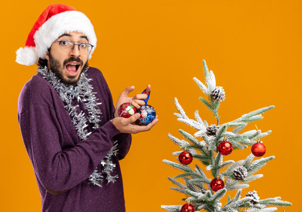 附近站在圣诞树旁的年轻帅哥戴着圣诞帽 脖子上戴着花环 手里拿着橙色背景上孤立的圣诞球帽子惊讶树