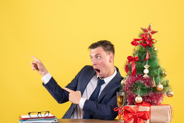 办公室前视图惊讶的人指着坐在圣诞树旁边的桌子上的东西和黄色墙上的礼物商人惊奇礼物