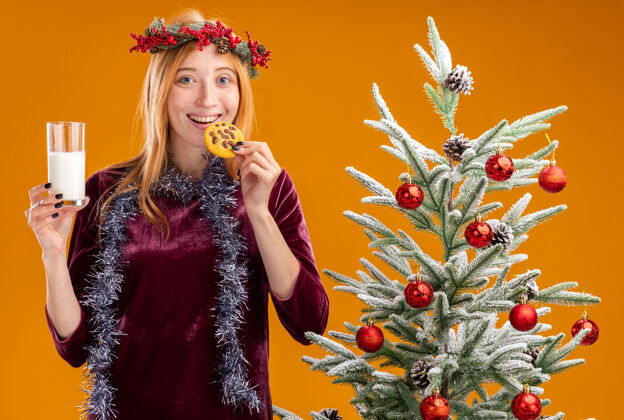 抱着微笑着站在圣诞树旁的年轻漂亮女孩 身穿红色连衣裙 脖子上戴着花环 手里拿着一杯牛奶 品尝着橘黄色背景的饼干杯子站着裙子