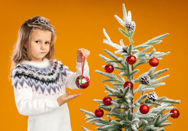 举行想着站在圣诞树旁戴着头饰的小女孩 脖子上戴着花环 指着孤立在橙色背景上的圣诞球圣诞节头饰思考