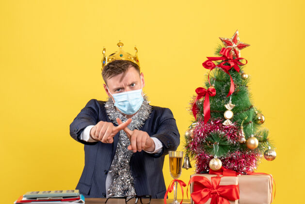 新商务人士交叉手指坐在圣诞树旁的桌子前 黄色墙壁上的礼物正常手指医疗面具