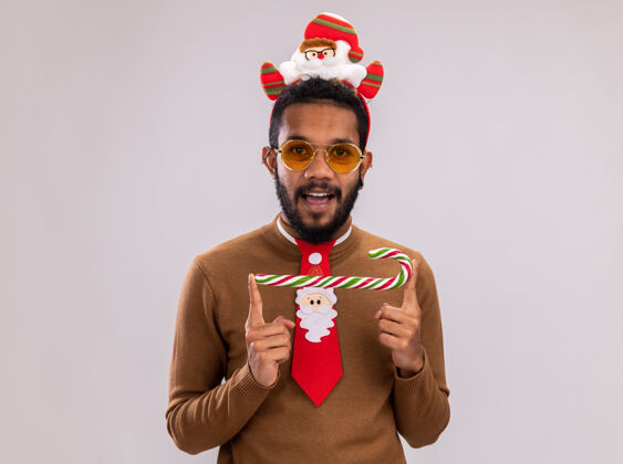 头一个身穿棕色毛衣 头戴圣诞老人耳环 打着有趣的红色领带 手持糖果手杖 站在白色背景下 看着摄像机 他开心地微笑着圣诞老人站着高兴