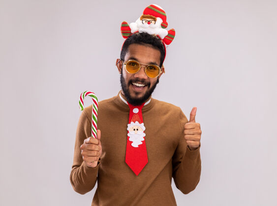 圣诞老人快乐的非洲裔美国人身穿棕色毛衣 头戴圣诞老人戒指 打着有趣的红色领带 手持糖果手杖 看着镜头 兴高采烈地微笑着 站在白色背景上竖起大拇指拇指糖果高兴