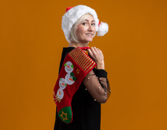 长袜微笑着的中年金发女人戴着圣诞帽站在侧视图中看着肩上拿着圣诞长袜的相机在橙色背景上隔离复制空间肩膀金发举行