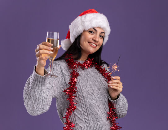 相机微笑着的年轻白人女孩戴着圣诞帽 脖子上戴着金箔花环 手里拿着节日火花和香槟 看着紫色背景上孤立的摄像机女孩帽子花环