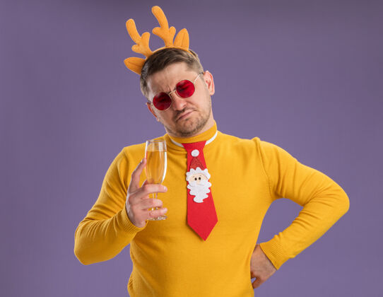 玻璃杯一个穿着黄色高领毛衣 戴着红色眼镜的年轻人 打着有趣的红色领带 戴着鹿角边 手里拿着一杯香槟酒 站在紫色的背景下 看着它不高兴眼镜搞笑站着