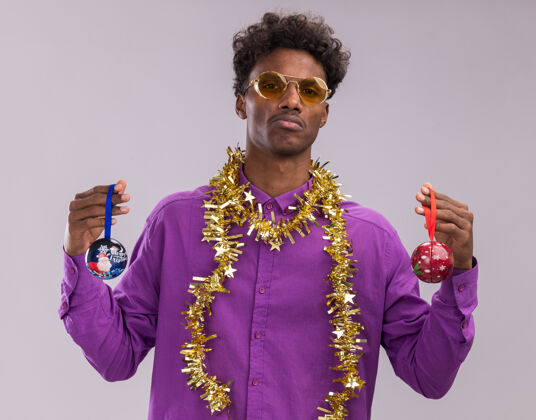 年轻不知所措的年轻美国黑人男子戴着眼镜 脖子上戴着金属丝花环 看着相机 手里拿着白色背景上孤立的圣诞饰品眼镜圣诞饰品周围