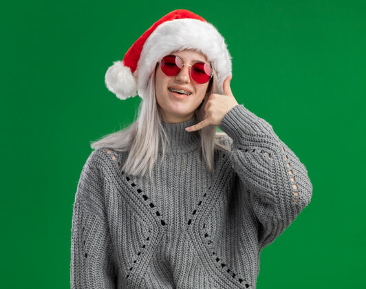 毛衣年轻的金发女郎穿着冬衣 戴着圣诞帽 戴着红眼镜 看着镜头 兴高采烈地站在绿色的背景上打电话给我年轻圣诞老人电话