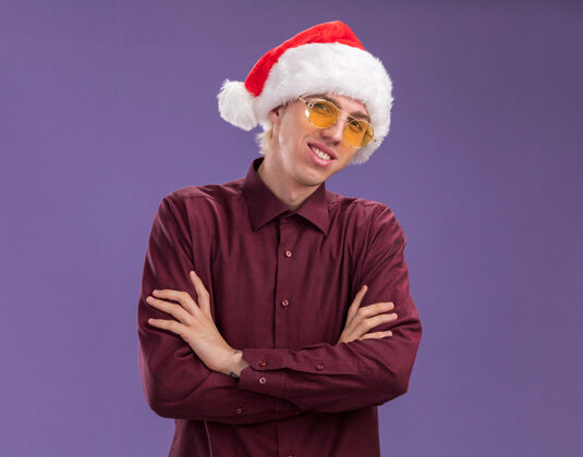 年轻微笑的年轻金发男子戴着圣诞帽 戴着眼镜 站在那里 以封闭的姿势看着紫色背景上有复制空间的相机姿势微笑闭上眼睛