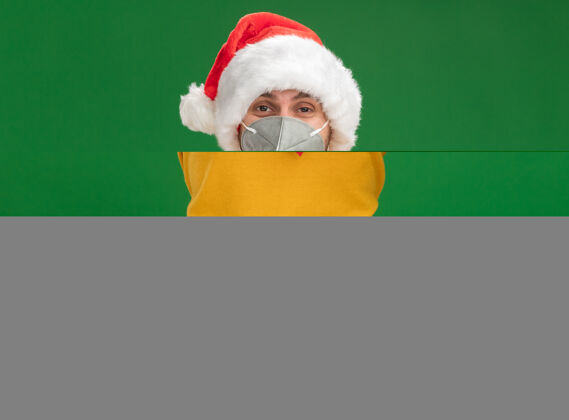高兴高兴的年轻白人男子戴着圣诞帽和领带 戴着防护面具 看着镜头做着心牌 隔离在绿色背景上圣诞节保护面具