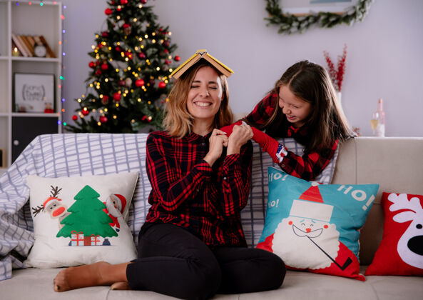 女儿微笑的女儿和母亲一起玩 头上抱着书 坐在沙发上 在家里享受圣诞节时光享受时间沙发