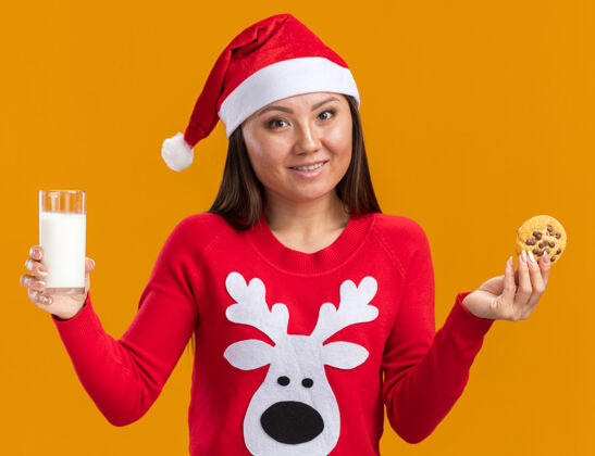 毛衣笑容满面的年轻亚洲女孩戴着圣诞帽 穿着毛衣 手里拿着一杯牛奶和饼干 背景是橙色的女孩拿着帽子