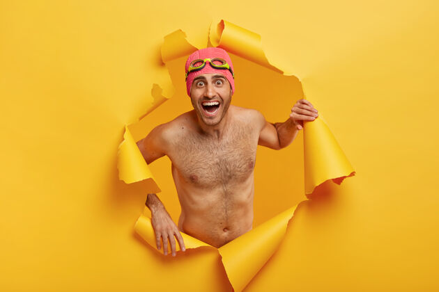 情绪化过度运动的男子游泳运动员赤膊上阵 戴泳帽和护目镜 在撕破的纸洞里摆姿势 开心地笑放松夏季高兴