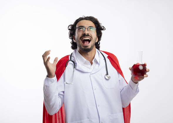 制服一个戴着眼镜 身穿医生制服 披着红色斗篷 脖子上戴着听诊器的快乐白人年轻人举起手站在玻璃瓶中 手里拿着红色的化学液体眼镜听诊器年轻