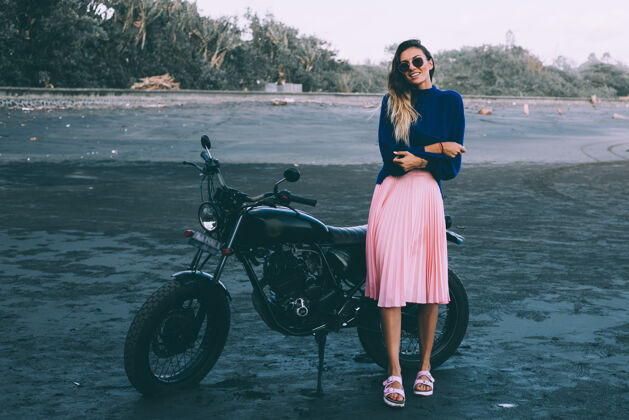 女孩自行车戴着太阳镜 穿着蓝色毛衣和粉色裙子的时髦女人的时尚肖像 站在黑色沙滩上的黑色自行车旁骑自行车的女孩坐着海洋