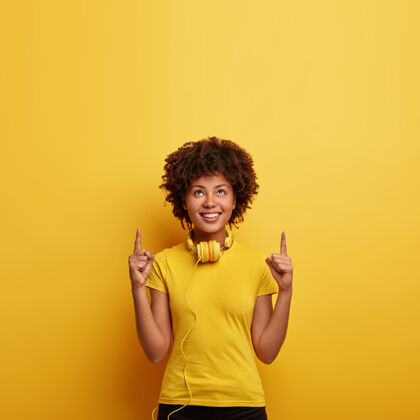 音频微笑的时髦女性用食指指向上方的照片 显示楼上的好地方 用耳机听最喜欢的曲目 穿着同一色调的亮黄色t恤与墙壁积极单色指向