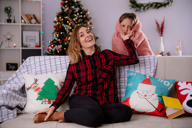 家微笑的母亲坐在沙发上 抱着女儿们裹在毯子里的脸颊 在家里享受圣诞节时光微笑毯子沙发