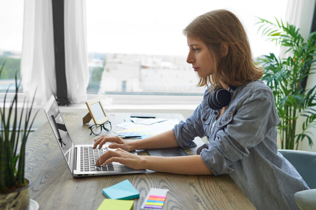 企业家人 现代科技和工作理念侧视图认真专注的年轻女性自由职业者戴着耳机在她的脖子上键盘上的便携式电脑 坐在窗户旁的木制办公桌坐着休闲女商人