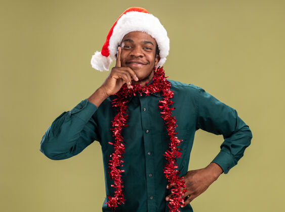 微笑戴着圣诞帽 戴着花环的非洲裔美国人站在绿色背景下 面带微笑地看着摄像机非洲人美国人帽子
