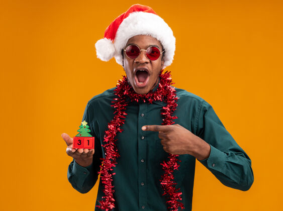 太阳镜戴着圣诞帽 戴着花环 戴着太阳镜 拿着印有新年日期的玩具立方体的非裔美国人站在橙色背景下 惊讶地用食指指着它立方体惊讶年