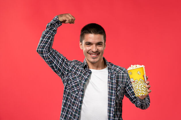电影正面图年轻男子拿着爆米花包 微笑着弯着腰站在浅红的墙上电影院电影男男孩光年轻剧院