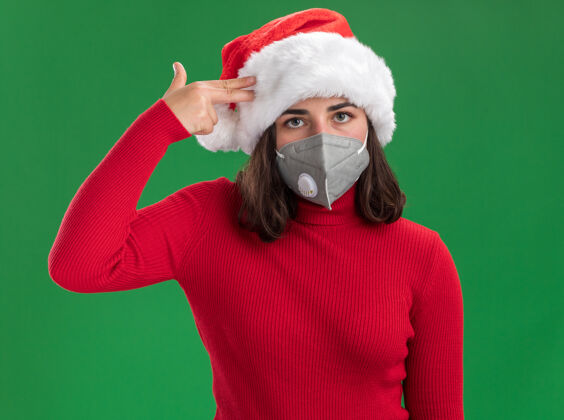 年轻穿着红色毛衣 戴着圣诞老人帽 戴着护面面具的年轻女孩用手指在太阳穴上做着手势 站在绿色的背景下 看着镜头 又累又无聊毛衣面具无聊
