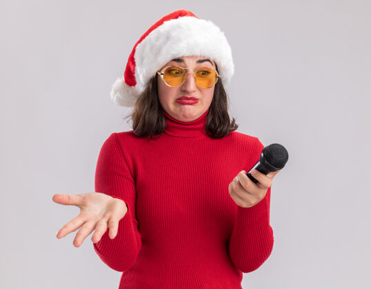 迷惑穿着红色毛衣 戴着圣诞帽 戴着眼镜 拿着麦克风的年轻女孩站在白色的背景上 用困惑的表情看着它眼镜毛衣女孩