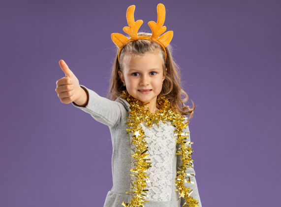 头发可爱的小女孩戴着圣诞发箍 脖子上戴着花环 在蓝色背景上孤立地竖起大拇指拇指显示女孩