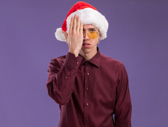 帽子年轻的金发男子戴着圣诞帽 戴着眼镜 看着相机遮住半张脸 手被隔离在紫色背景上 留有复印空间年轻金发脸
