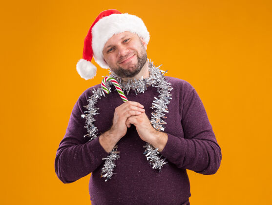 圣诞老人高兴的中年男子戴着圣诞帽 脖子上戴着金箔花环 手里拿着圣诞甜手杖 看着隔离在橙色背景上的相机周围中年圣诞节