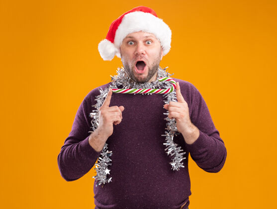 脖子惊讶的中年男子戴着圣诞帽 脖子上戴着金属丝花环 手握圣诞甜手杖 水平地看着隔离在橙色背景上的相机圣诞节中年手杖