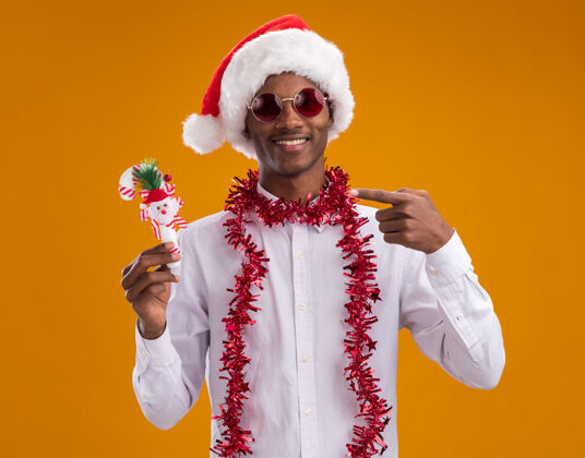 圣诞老人微笑着的年轻的非洲裔美国人戴着圣诞帽 戴着一副眼镜 脖子上戴着金箔花环 拿着并指着糖果手杖饰品 看着孤立在橙色背景上的相机手杖周围年轻