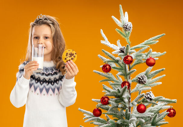 圣诞快乐高兴的小女孩站在圣诞树旁 戴着头饰 脖子上戴着花环 手里拿着一杯牛奶和饼干 背景是橙色的头饰快乐拿着