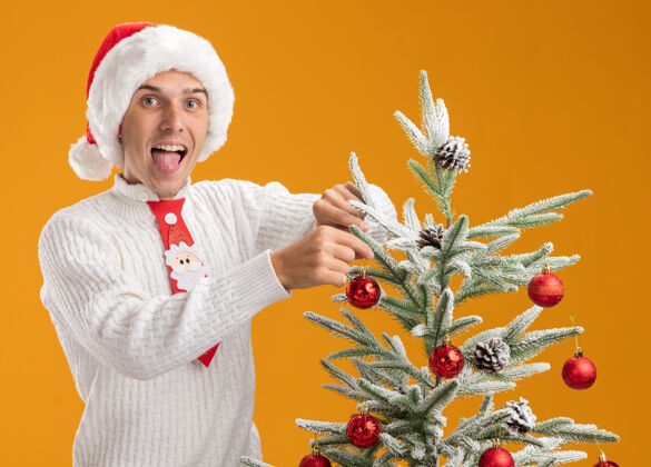 年轻可爱的年轻帅哥戴着圣诞帽打着圣诞领带站在圣诞树旁用圣诞球装饰圣诞树展示装饰穿着