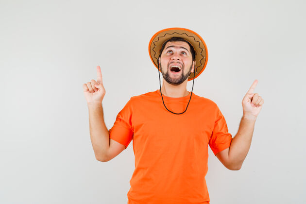 模特年轻人指着手指在橙色的t恤衫 帽子和看起来高兴 前视图人帽子衬衫