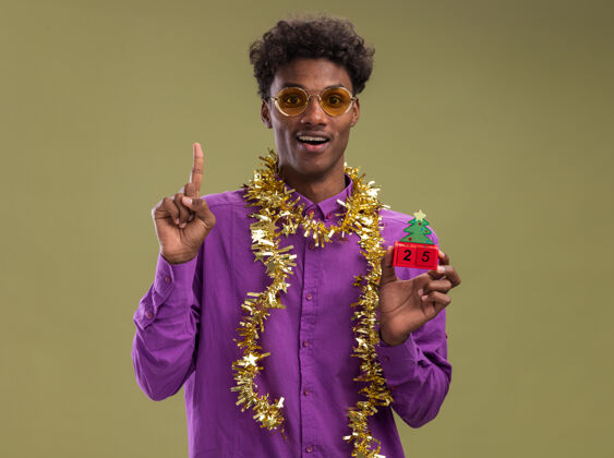 非裔美国人令人印象深刻的年轻非洲裔美国人戴着眼镜 脖子上戴着金属丝花环 手里拿着圣诞树玩具和日期 看着相机指着橄榄绿背景上的孤立橄榄印象深刻圣诞节