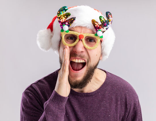 情绪化一个穿着紫色毛衣 戴着圣诞帽 戴着滑稽眼镜的年轻人站在白色背景下看着摄像机 情绪激动地大叫着搞笑圣诞老人站着