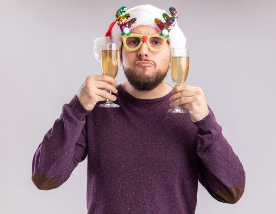 圣诞老人年轻人穿着紫色毛衣 戴着圣诞帽 戴着滑稽的眼镜 手里拿着两杯香槟 歪歪着嘴 站在白色背景下的新年假期概念搞笑帽子眼镜