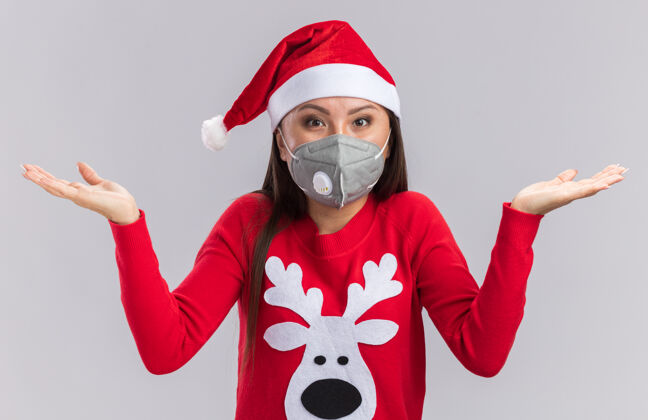 传播惊讶的年轻亚洲女孩戴着圣诞帽 穿着毛衣 戴着医用口罩 双手被隔离在白色背景上亚洲人毛衣医学