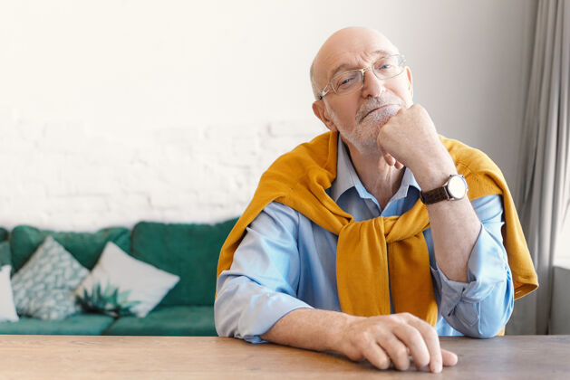 老人戴着长方形眼镜 肩上系着毛衣的资深秃头心理学家坐在家里办公室的空木桌旁 等待着他的委托人 有着若有所思的表情祖父眼镜桌子