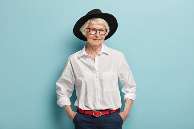 老年人时尚成熟的女人戴着时髦的黑帽子 白衬衫 红腰带裤子 手放在口袋里 表情欢快 隔着蓝色的墙人 年龄 时尚的概念女人裤子皱纹