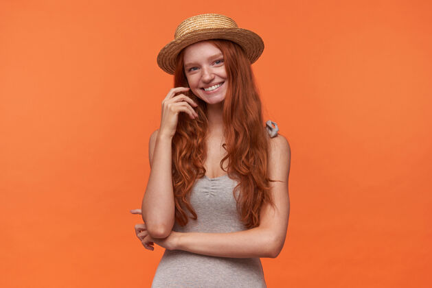 牙齿摄影棚拍摄的年轻红发美女穿着休闲服 头戴水手帽 站在橙色背景下 面带迷人的微笑看着镜头 抚摸着她波浪形的头发稻草情绪长