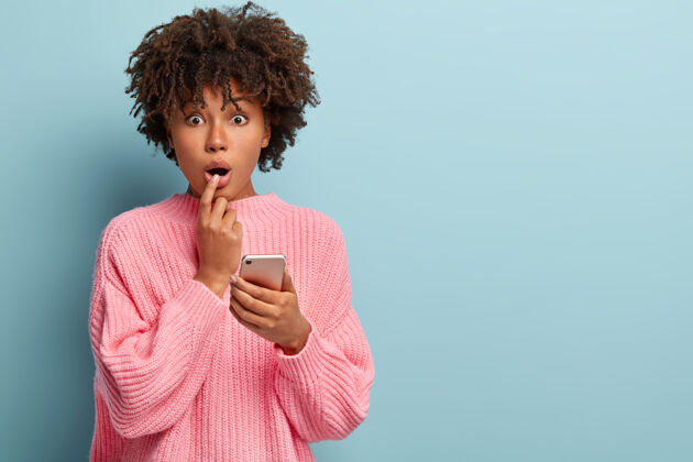 短信惊讶的美国黑人女性的照片在网上购物 拿着手机 垂头丧气 表达惊奇 穿着宽松的针织套头衫 收到朋友的奇怪通知非洲理发非洲