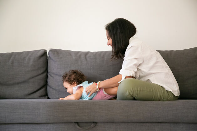女人快乐的黑发妈妈抱着可爱的小女儿在灰色沙发上侧视图父母和童年的概念视图白种人侧