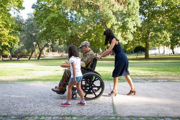 父母微笑的军人和家人在城市公园散步长发的母亲推着轮椅小女孩和残疾的父亲散步和交谈家庭户外 周末和残疾概念孩子轮椅妈妈