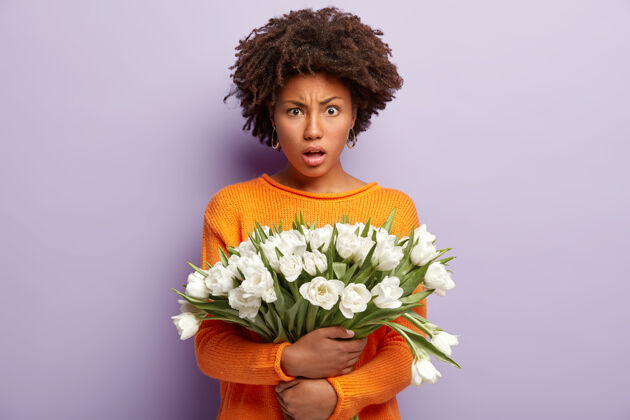 不满愤愤不平的女人生气地看着 拿着白色的花 穿着橙色的休闲套头衫 在紫色的墙上做模特 表达负面情绪 听到坏消息郁金香女人惊讶花束困惑