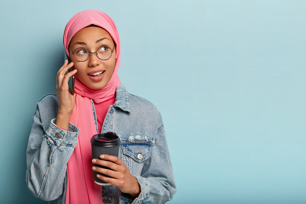 手机欣喜的阿拉伯女人肖像通过手机整理 捧着一杯咖啡 收到好报 侧目交谈头巾远离
